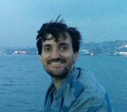 Smiling white male with scraggly moustache and beard and windswept hair with the Turkish strait behind