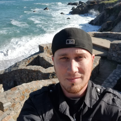 White man with black backwards baseball cap in front of rocky beach