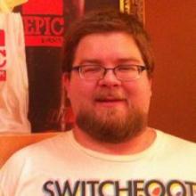 A rotund, bearded man with short, brown hair wearing a colorful T-shirt.