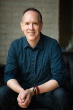 photo of Dan Moriarty, wearing dark blue shirt, sitting, with brick wall as backdrop