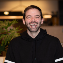 Eric Leech, miling broadly while wearing a black hoodie, standing indoors with blurred background elements behind