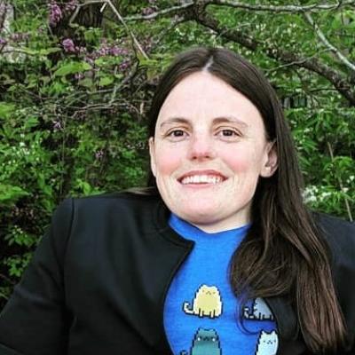 headshot of woman with long brown hair, blue a11y cats shirt and black blazer