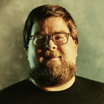 The head of rotund, bearded man with short, brown hair and glasses lit mostly from the right side.