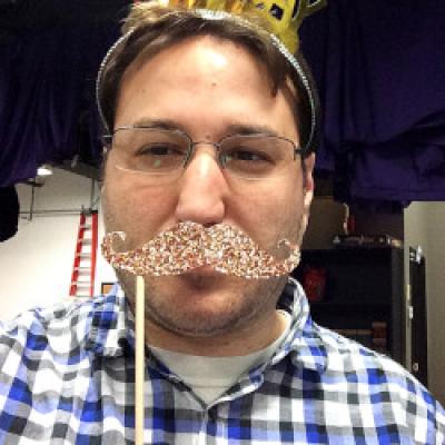 Matthew hiding behind a glitter moustache masquerade with a plastic tiara wearing a blue, gray and white plaid shirt.